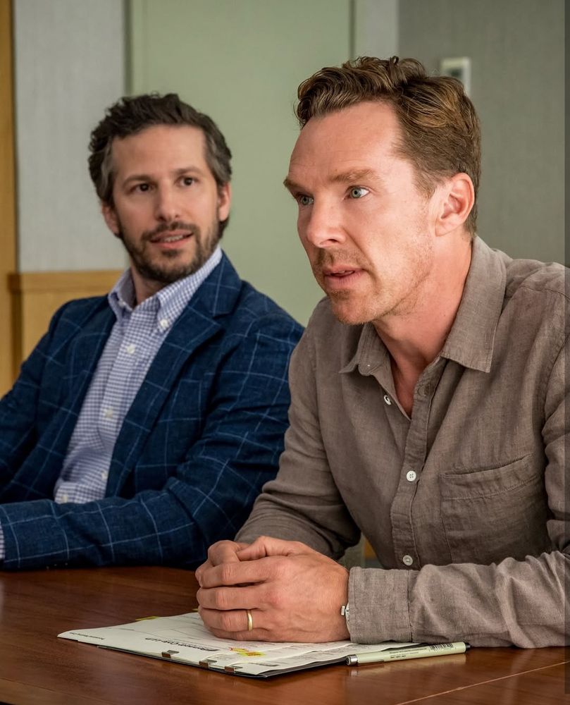Os atores Andy Samberg e Benedict Cumberbatch em imagem do filme The Roses. Ambos estão sentados à mesa, Benedict em primeiro plano de camisa acinzentada, corpo inclinado para a frente, antebraços apoiados na mesa. Andy ao lado dele de camisa e paletó quadriculados em tons de azul, olha para Benedict.