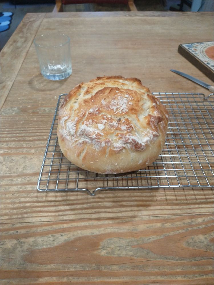 A round loaf of bread sits on a metal cooling rack on a wooden table with an empty glass to the left and a knife to the right.