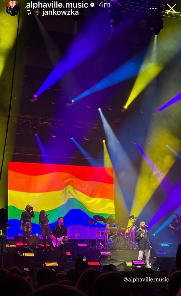 An Insta story reel picture showing Alphaville on stage in front of a screen with a giant rainbow flag on it