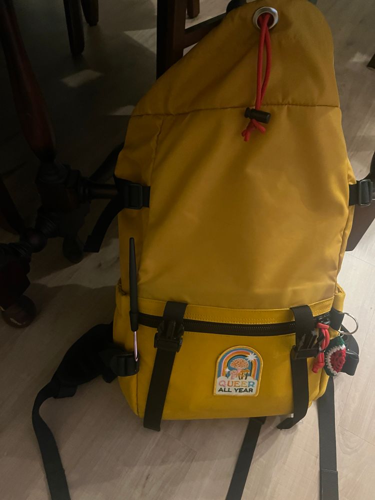 A yellow backpack on a light wood floor black straps and a “queer all year patch” 
Hinting at the found crochet hook
