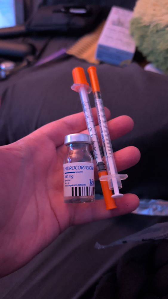 A vial of injectable hydrocortisone next to two syringes held on my own small hand. They're all empty, because they've been pulled up and used. 
