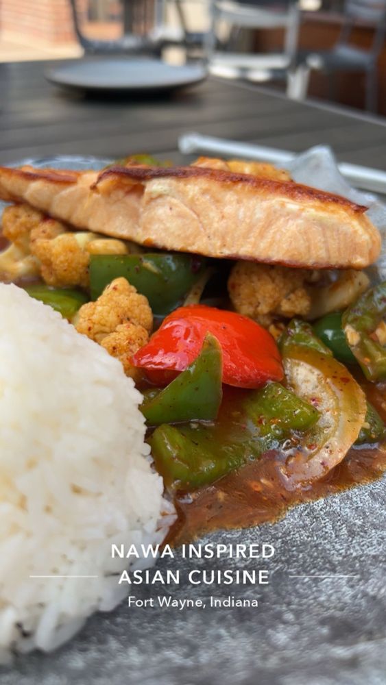 In the foreground, there’s a perfectly domed serving of white rice. Nestled beside it is a medley of colorful vegetables—green and red bell peppers, tender cauliflower florets, and sliced onions—coated in a glossy, rich, spicy-sweet Kung Pao sauce, flecked with chili and seasoning.

Atop the vegetables sits a generous fillet of salmon, seared to a beautiful golden brown with a crisp, caramelized exterior, hinting at its tender and moist interior. The dish is presented on a stylish ceramic plate, with an inviting outdoor dining setting softly blurred in the background.

snap caption: NAWA Inspired Asian Cuisine Fort Wayne, Indiana