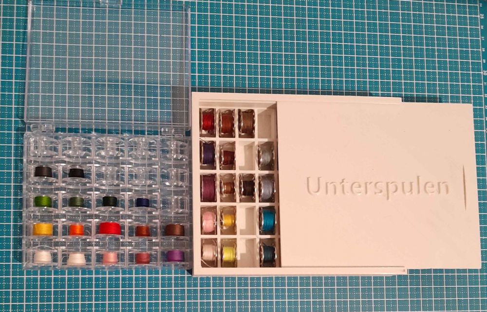Two bobbin cases the smaller one is store bought with plastic bobbins
The bobbins are sorted by thread color. 
The larger white case is 3D printed and holds some metall bobbins. The lid is partly opend and spells out "Unterspulen" ontop