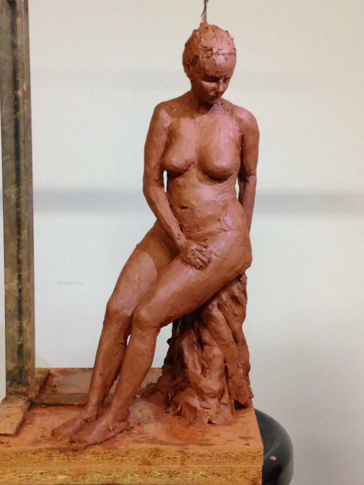 Escultura en barro de mujer sentada en una banqueta con las piernas juntas y mirando a un lado y abajo.