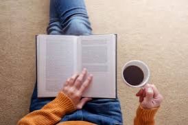 Photo of an open book on a woman's lap. She is holding a coffee and reading.