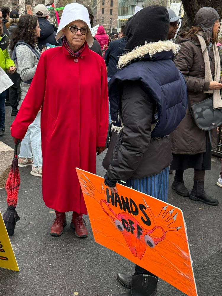 At the Hands Off march, NYC April 5, 2025, a woman dressed as a character from A Handmaid's Tale talks with someone holding a sign with a uterus that says Hands Off