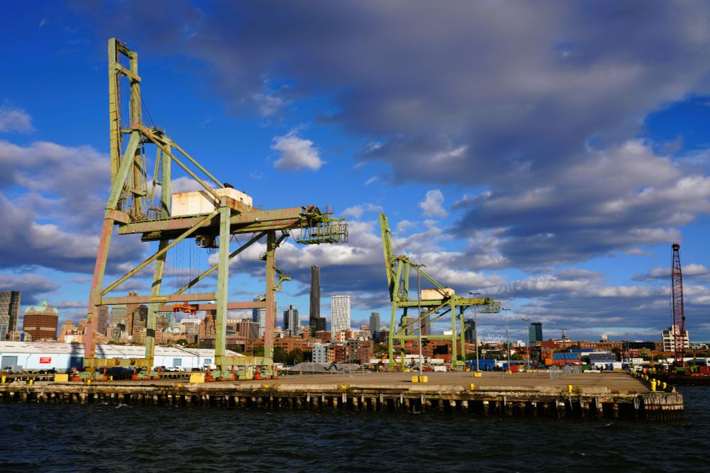 Giant green cranes resemble chairs in the cargo container terminal in Brooklyn. The sky is blue with soft clouds. The cranes are used in ports to load and unload cargo, primarily shipping containers, from vessels.
