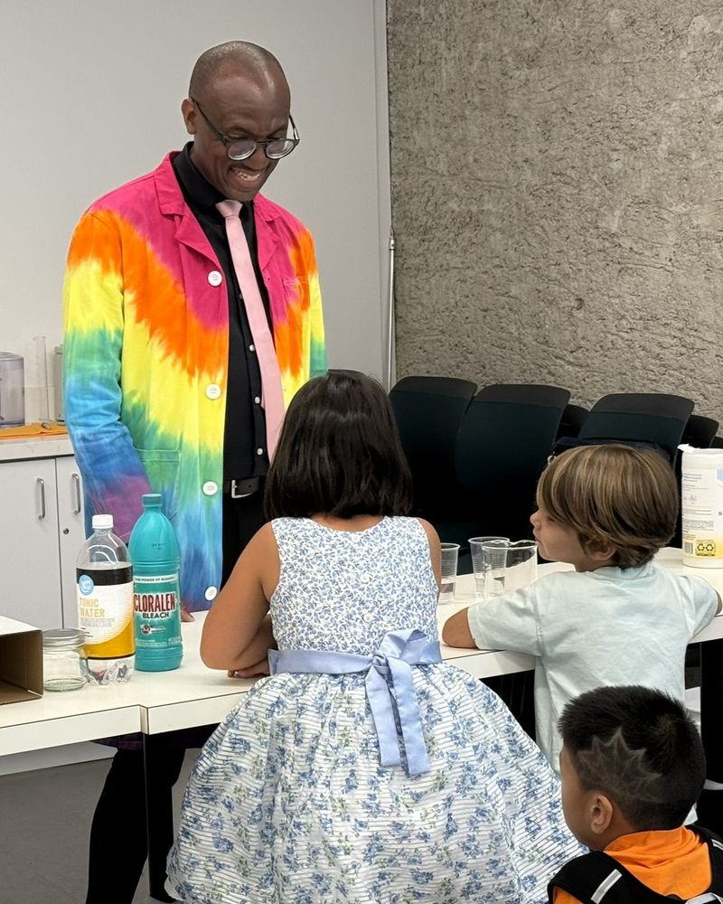 Dr. André is wearing a rainbow lab coat and pink tie and talking to 3 children at the American Museum of Natural History. One of the demonstrations he did that showcase chemistry in action was the 
Briggs-Rauscher: Oscillating Clock
Three colorless solutions are combined into a large beaker and the mixture becomes amber, then blue-black, and then colorless again. This sequence of color changes repeats!
Colorless: I2 is consumed
Amber: The amber color results from the production of I2
Blue: The deep blue color is the result of the I- and I2 binding to starch in the solution.
