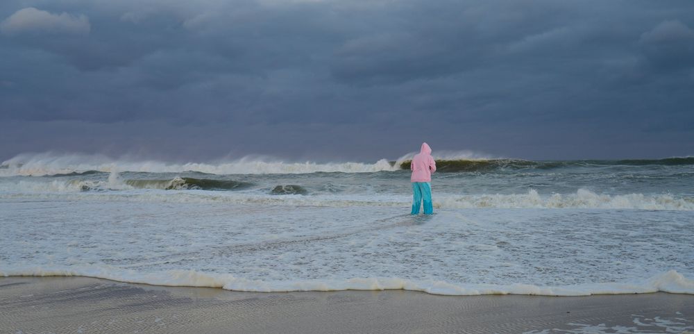 A person stands in the water. There are huge waves in front of them, the sky is dark and cloudy.