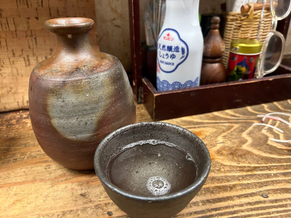 緋襷のような赤い焼き色、焦茶色の肌の備前焼きのとっくりに燗酒。
やはり備前焼きの黒いぐい呑みに、泡の残る燗酒が入っている。