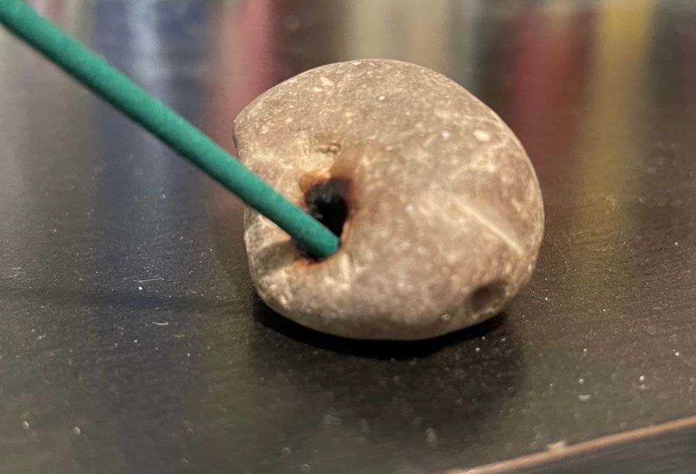 A small grey rock with a hole which is perfectly sized to hold a stick of incense. To demonstrate this, it is holding a stick of incense. 