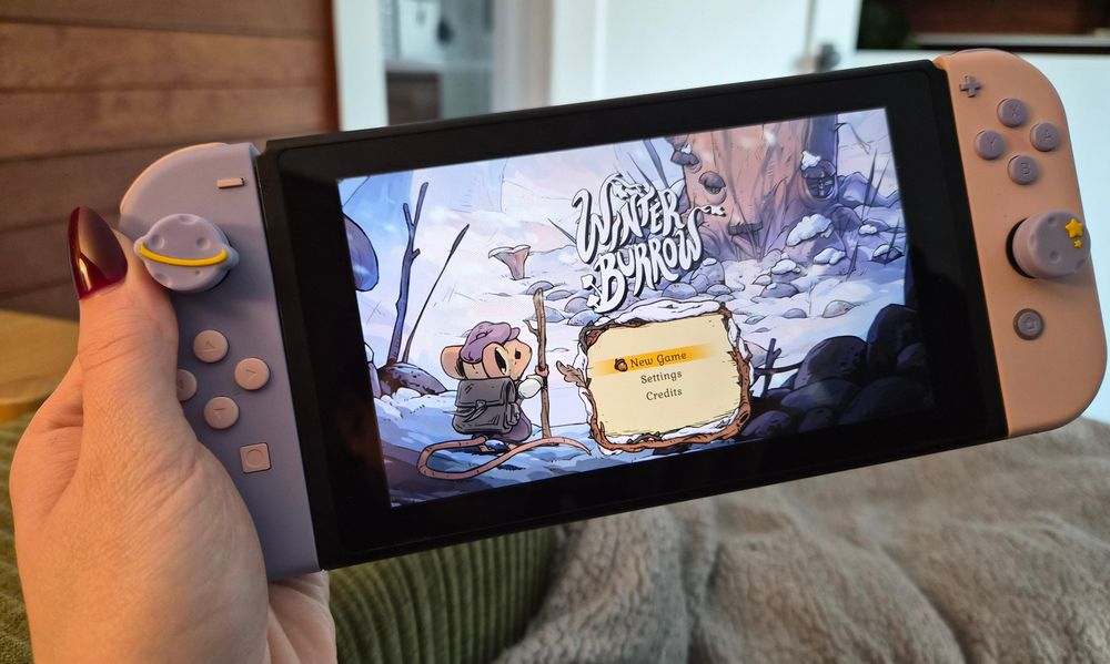 holding a purple and pink switch with the start screen for Winter Burrow. the screen shows a cute illustrated mouse looking out into a wintery forest 