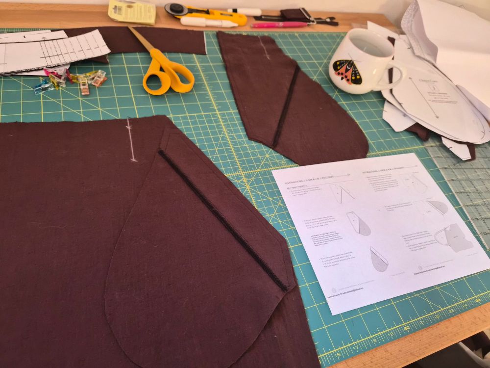 sewing project ephemera spread over a table, including brown fabric marked with chalk, a sheet of instructions, cup of coffee, and various clips and scissors.