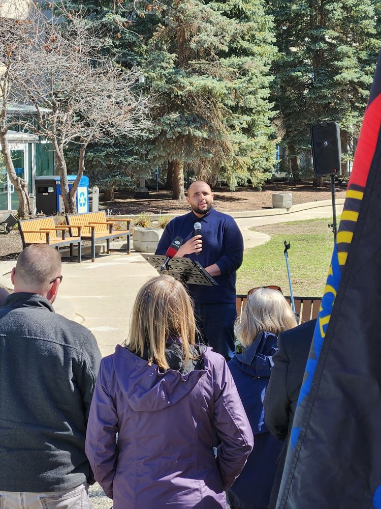 Photo of Calgary City Councillor Courtney Walcott, a medium-skinned man with a shaved head and a beard, standing at a podium in a park on a sunny day addressing a crowd with a flag in the foreground.
