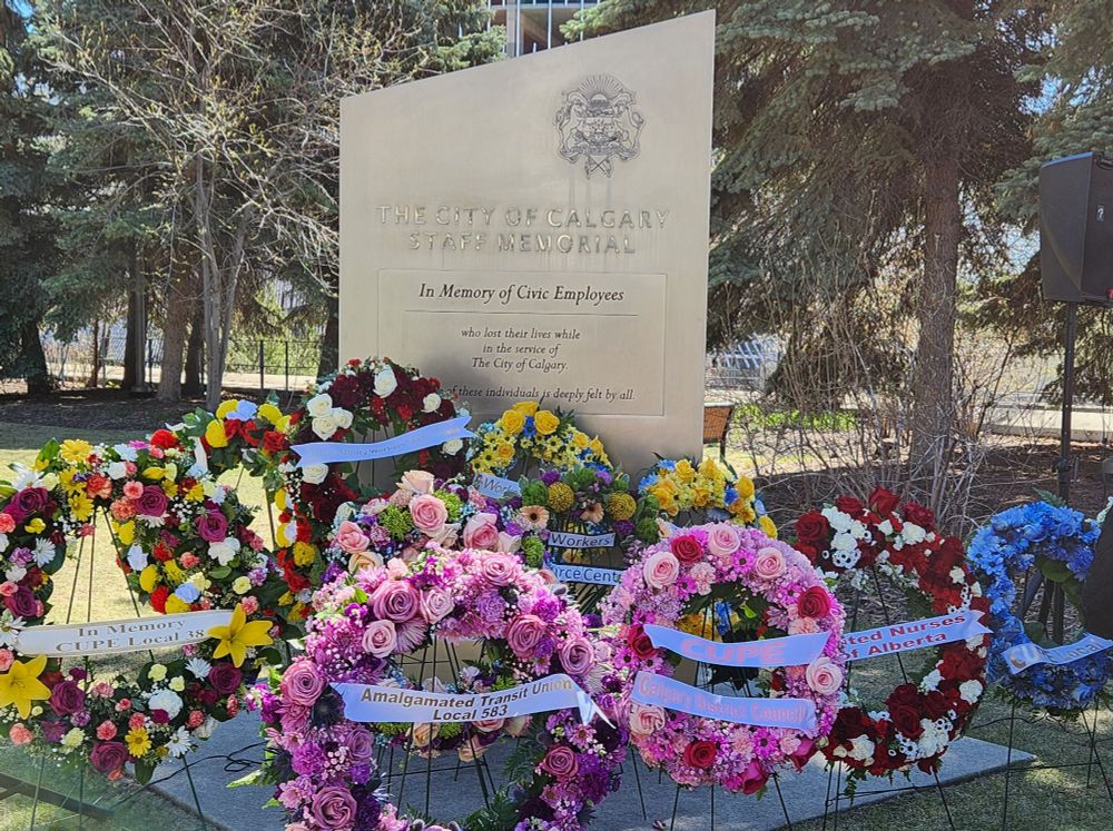 Photo of the City of Calgary Staff Memorial monument, a silver rectangular-shaped monument with the city seal at the top. The monument is surrounded by about a dozen wreaths adorned with various colours flowers - pink, purple, red, light blue, white and yellow.