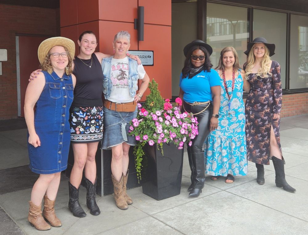 Photo of six women standing around a planter with pink flowers outside an office building in Calgary, Alberta. They are all wearing Stampede outfits - some with denim, cowboy boots, country-style embroidery and hats. 