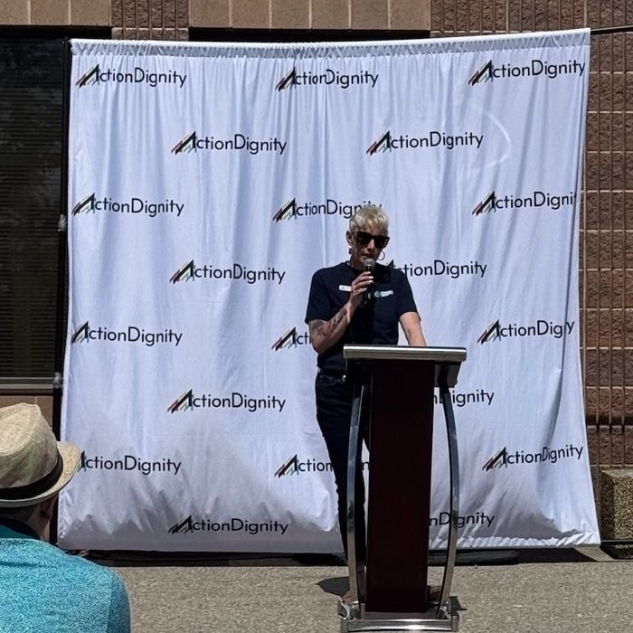Photo of a woman dressed in a black t-shirt, black pants and sunglasses with short, blonde hair. She is standing at a podium in front of a backdrop with the ActionDignity logo holding a microphone addressing a crowd.


