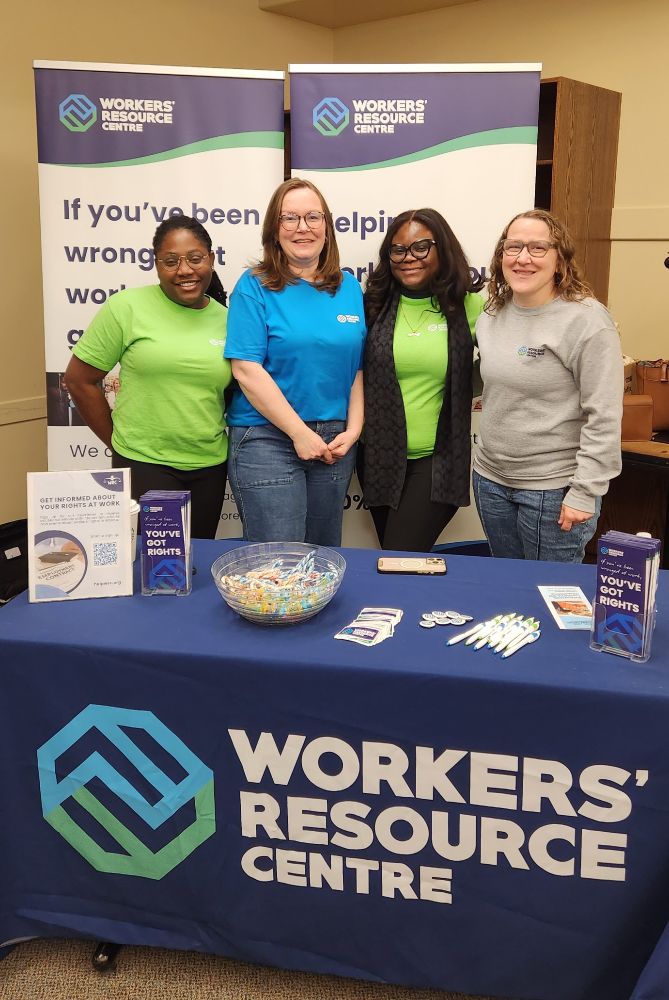 Photo of four bespectacled, long-haired women standing side by side behind a table labeled "Workers' Resource Centre," with brochures, pens, stickers and candy. There are two dark-skinned women in bright green t-shirts with white Workers' Resource Centre logos, and and two light skinned women, one who is wearing a bright blue shirt and the other a light grey sweater with the Workers' Resource Centre logos on them.