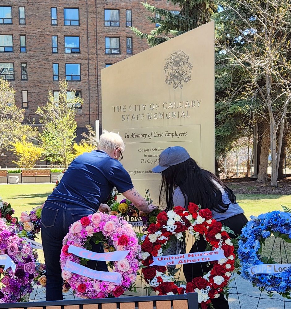 Photo of two women, one tall with  short, white-blonde hair and the other a little shorter with long straight black hair and a ball cap, bending down to lay a wreath in commemoration of Alberta workers who died at the City of Calgary Staff Memorial monument, among about a half dozen other bright wreaths with pink, blue, red and yellow flowers.