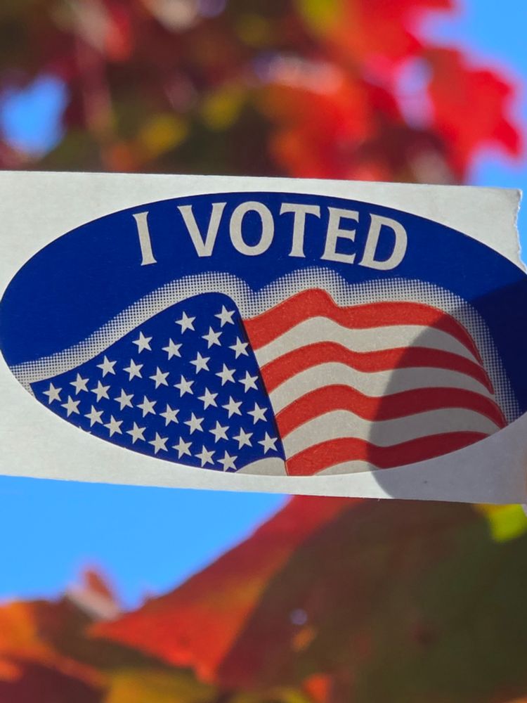 A classic oval "I Voted" American flag sticker still attached to its backing is in focus in the center. Out of focus in the background are red and green autumn leaves against a bright blue sky.