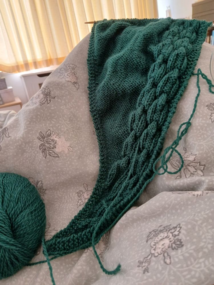 A shawl in progress, in rich green, with cables forming two chains up the right hand side. The piece is currently around 40cm long.