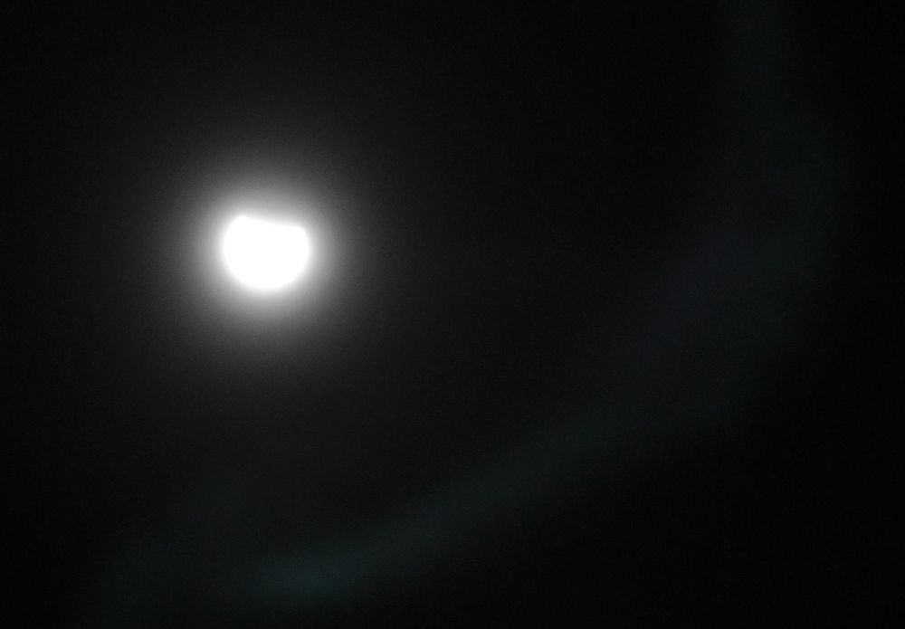 Overexposed shot of today's eclipse.
A dark background with a bright white misshapen blob and a swirl of cloud to the bottom right.