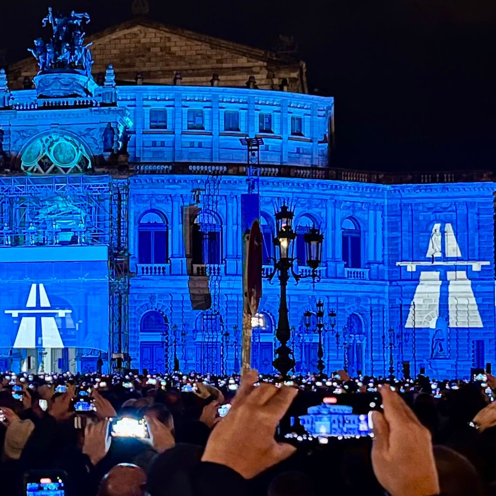 Kraftwerk live in Dresden. Projektionen tauchen die Semper-Oper in blaues Licht. Weiße Autobahn-Logos sind groß zu sehen. 
