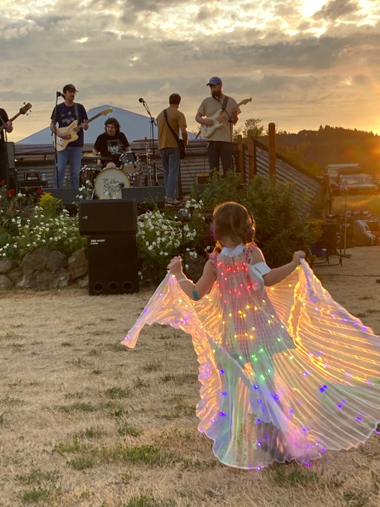 A child wearing a diaphanous cape with rainbow LED lights in it dances in front of a 5-piece rock band playing on the deck of an ancient pickup truck. In a field with the sun setting behind.