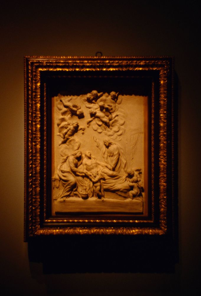 A relief sculpture in a golden frame, seemingly depicting two figures aiding a third who is reclining, possibly sick or injured