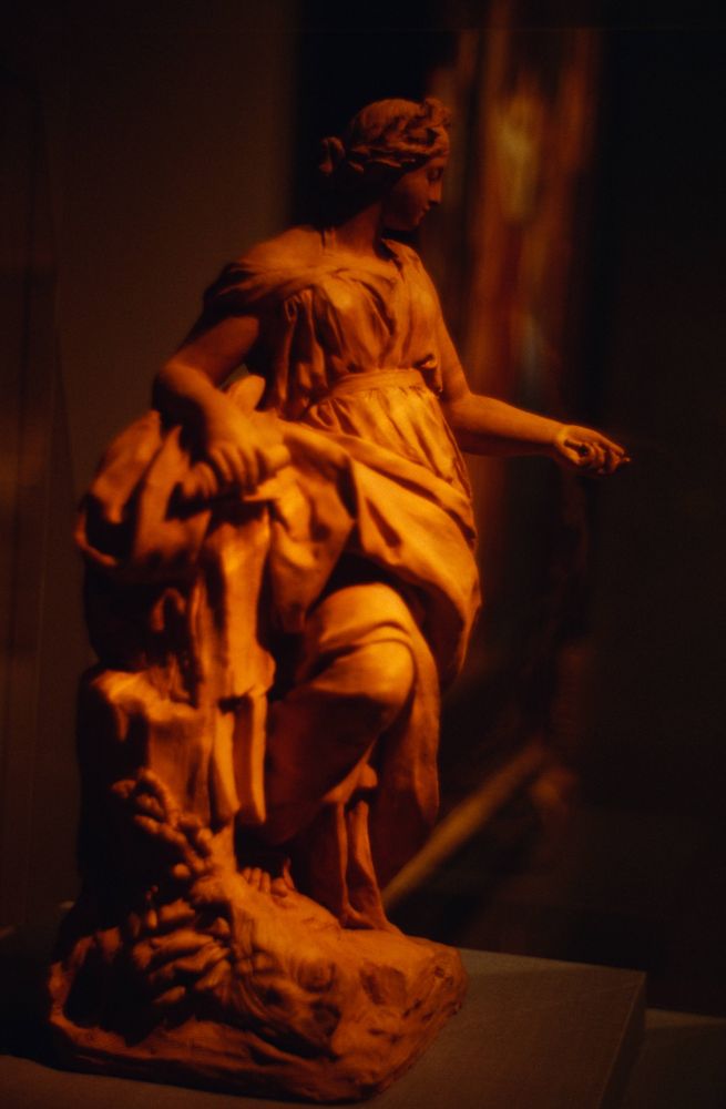 A very orange-cast sculpture of a woman with a basket in her right arm, looking at her left hand held in front of her. Very moody