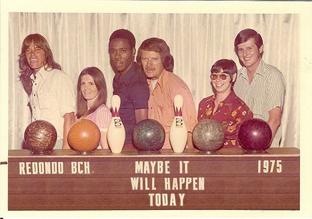 A time-aged photo of a bowling team posing behind a display of bowling balls and pins. From left to right, we've got a long-haired white guy making a 'hell yeah!' face and probably hearing REO Speedwagon in his head. Then there's a pretty girl with straight long hair in a pink shirt very consciously doing a posing for a photo face. Then a black guy in a blue polo to prove everyone here is cool, next to the oldest dude on the team with longer hair and a 'stache in a peach-colored bowling shirt. Then a girl with a cute bob and sunglasses in a floral shirt, and finally there's Ed who there isn't a lot to say about. Below the ball display is a wooden sign with tracks for placing movable plastic letters. In the top left it reads 'REDONDO BCH.' and top right is '1975' and between those is the mysterious phrase 'MAYBE IT WILL HAPPEN TODAY'.
