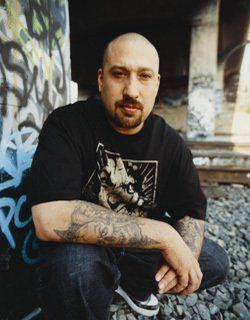 B-Real squatting down with arms rested on knees, wearing black t-shirt, jeans and with a bald head and goatee.  He is near railroad tracks and concrete covered in spray paint tags.