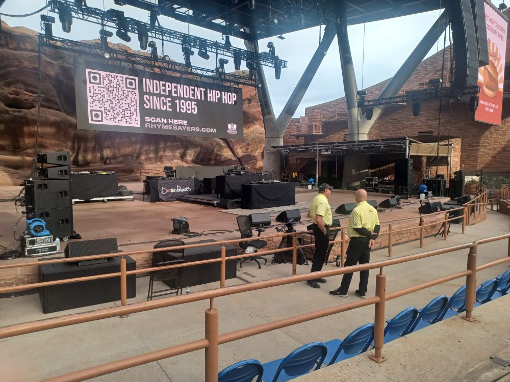 The pre-show stage at Red Rocks Amphitheater in Morrison, Colorado on September 19, 2025.  4 long tables with turntables, mixers and microphones are on stage.  Security stands in the foreground.
