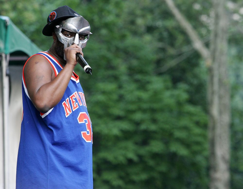 MF DOOM is a Black man on stage during a performance.  He wears a shiny silver gladiator style mask, black flat bill cap with red insignia turned sideways and a royal blue and orange New York Knicks jersey.  He holds a microphone to his mouth with his right hand.  There are trees with green leaves in the background.