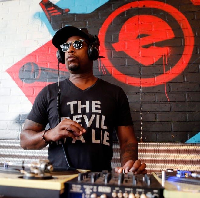 Talib Kweli is a Black man wearing dark sunglasses, a dark flat bill cap and black t-shirt that says "The Devil is a Lie" in white block letters.  He wears DJ headphones and has his left hand on a mixer situated between two vinyl turntables.  The brick wall behind him is painted with red and black artwork.