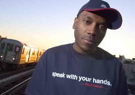 Rob Swift near the train tracks and a passing sun drenched train, wearing a navy blue & red baseball cap slightly turned to the side and navy blue t-shirt that reads in white lettering 'speak with your hands'