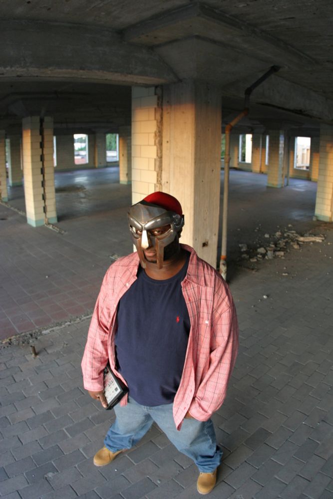 MF DOOM is a Black man wearing a shiny silver gladiator style mask, red cap, navy t-shirt under a red and white unbuttoned dress shirt, blue jeans and brown tims.  He stands in an empty parking garage with concrete pillars.