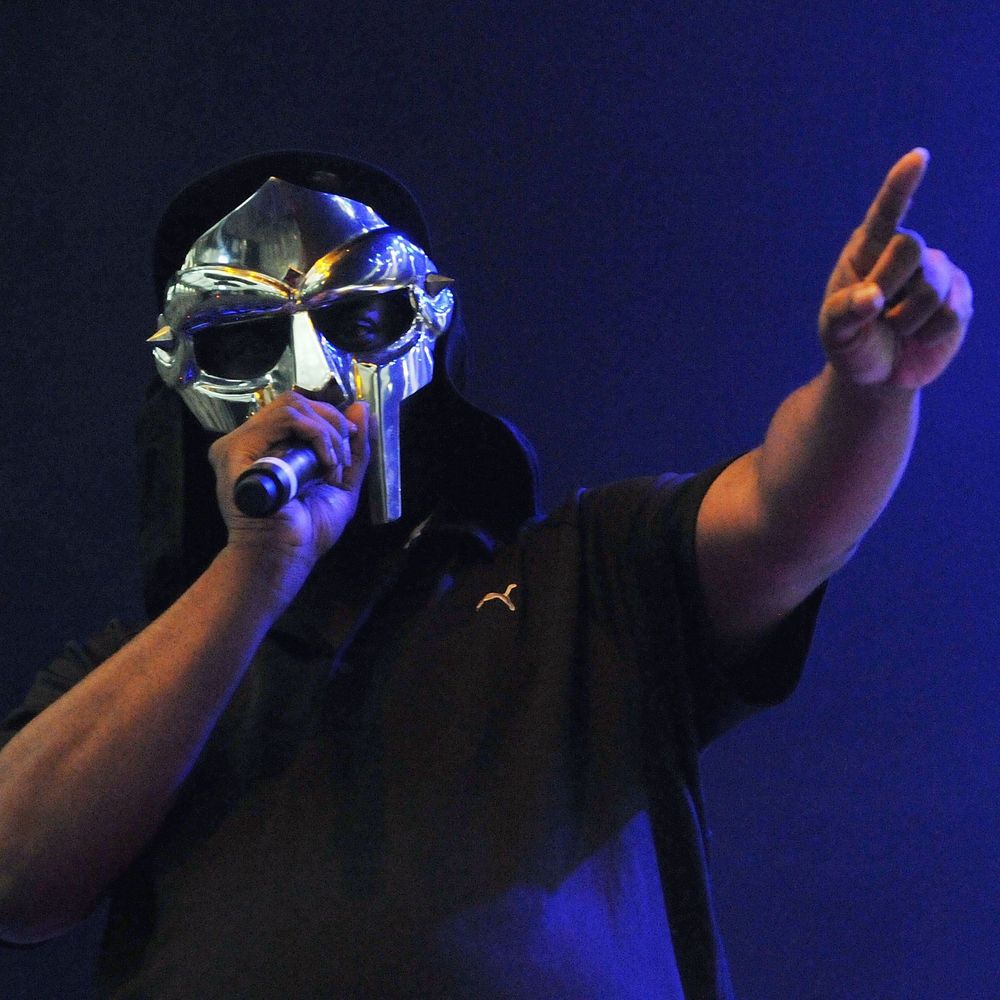 MF DOOM is a Black man with a shiny silver gladiator style mask.  He is holding a microphone to his mouth with his right hand and pointing his left index finger to the crowd during a live performance