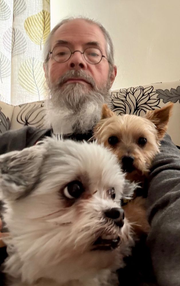 Elderly biblical scholar with two dogs in his lap, in the foreground the head of Flora, a white-haired Lhasa Apso/Maltese mix (whose jaw is offest to the right oif her nose, poor dear), and between Flora and the old man is Minke, a sandy brown-grey Yorkshire terrier, anxious, barky and annoying.