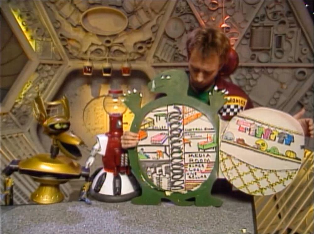 Scene from Mystery Science Theater 3000: Crow T. Robot and Tom Servo show off their oversized interactive model of Gamera to Joel. It is a big green turtle with a hinged shell that opens to a spiral staircase and four levels of different rooms.