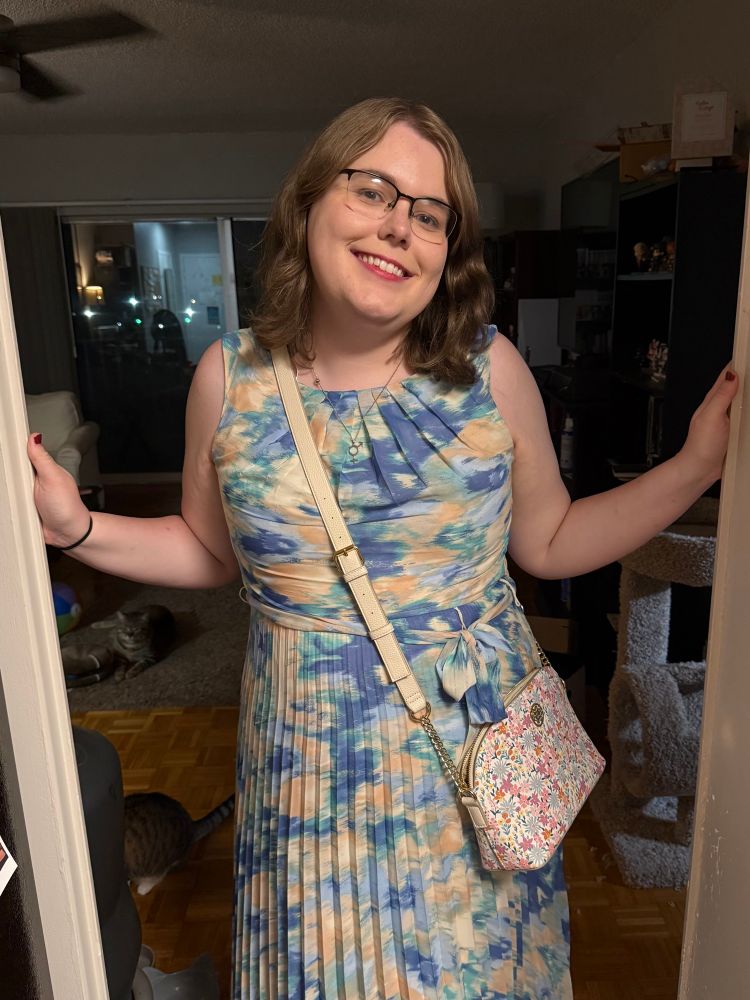 a gorgeous woman wearing a pretty dress of various blues and a tan color, smiling brightly in a doorframe