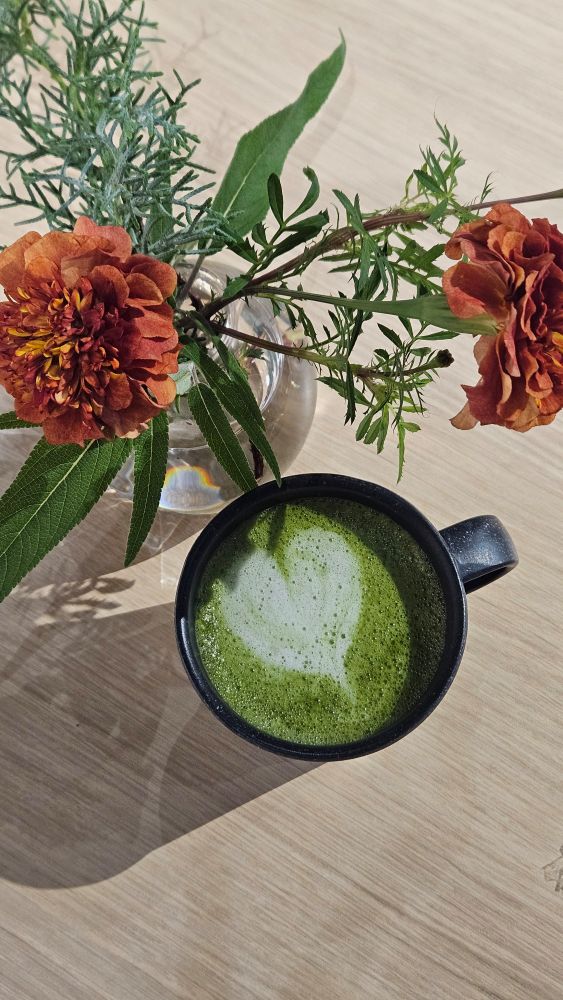Tasse de matcha latte avec un coeur dessiné en latte art. Le tasse est posée à côté d'un vase avec des fleurs.