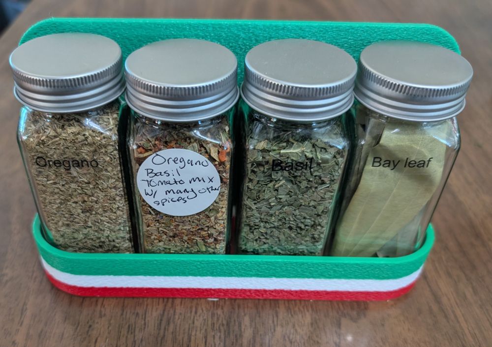 A 3d Printed Spice Rack. It is mostly green, and then there is a stripe of white and red, so it is similar to an Italian flag. The spice rack now contains a selection of spices common to Italian cooking.