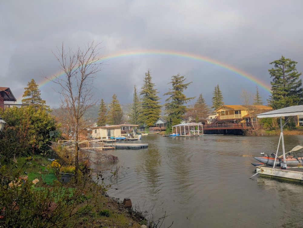 An image of a canal connected to Clearlake, CA. Several houses with docks are shown. The sky is dark, it is raining, and the main feature is a full rainbow bridging the canal.