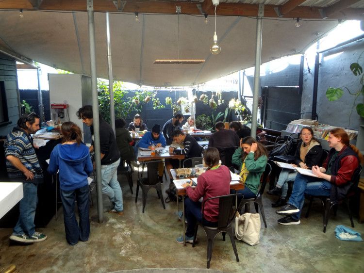 A group of people sitting at tables in the covered outdoor space behind Obz Books in Observatory, Cape Town. They are drawing comics on A4 sheets of paper.

Photograph: Mandy J Watson