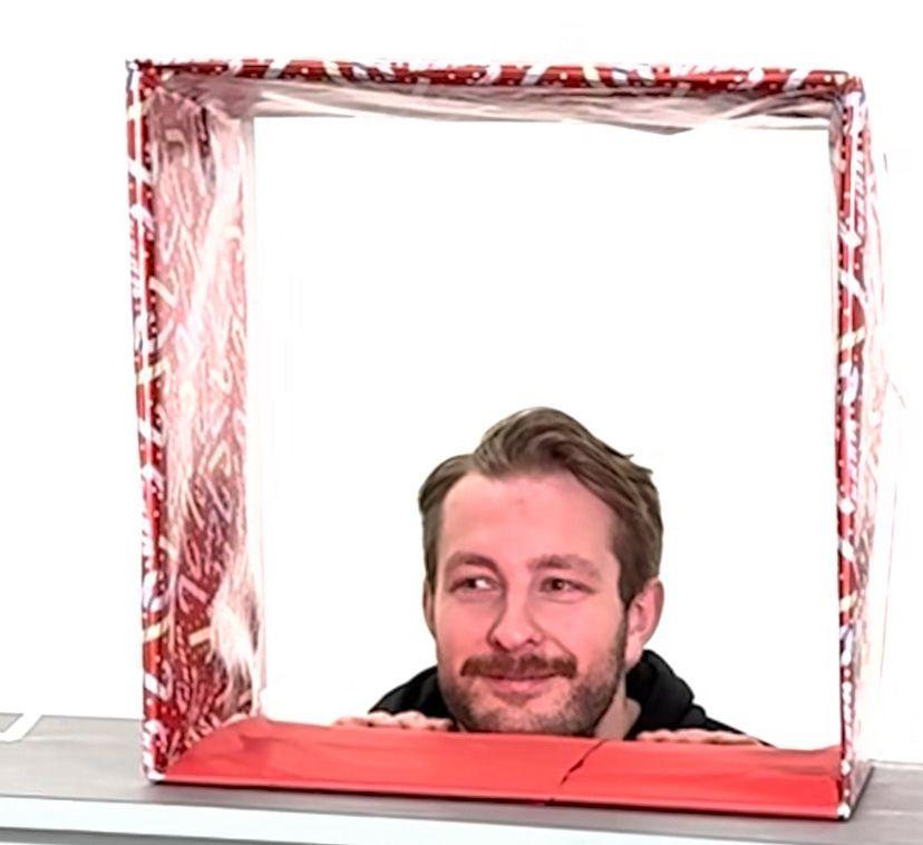A cardboard square wrapped in red, metallic, candy cane-patterned wrapping paper sits upright on a table. Linus Ullmark is kneeling behind the table, with only his head and hands visible, as he looks through the “present.” The background is a white wall. 