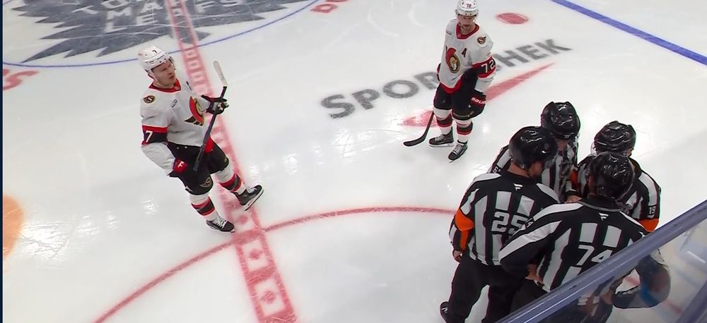 Brady Tkachuk and Thomas Chabot, wearing the Sens white away jerseys, stare at the officials as the four of them converse beside the boards. 