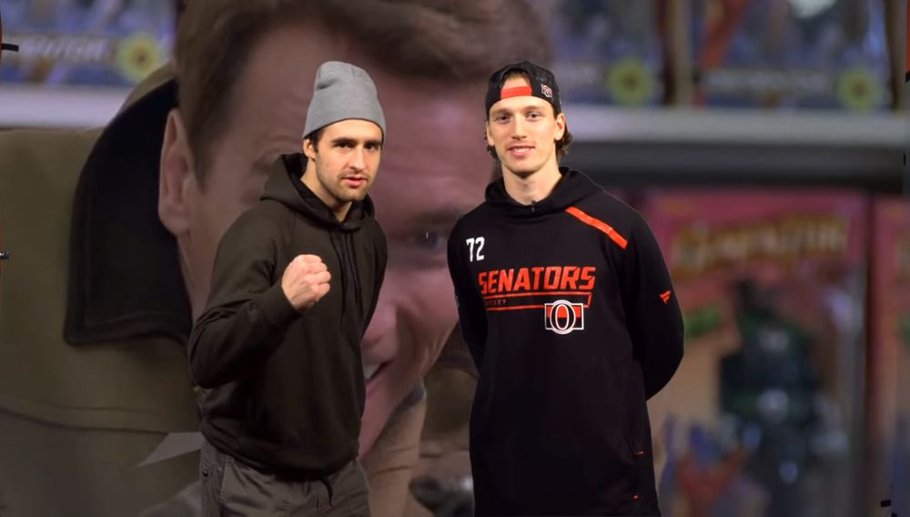 Colin White and Thomas Chabot stand in front of a scene from Jingle All the Way, with Arnold Schwarzenegger inside a toy store before Christmas. White is wearing a black hoodie, grey pants, a light grey beanie, and has his left arm up in front with a fist and his right arm behind him, with a piece of paper in his hand visible near his back. Chabot is wearing a black hoodie with “Senators” in red text, the Sens “O” logo underneath it on the right, and a 72 on his left shoulders, and a backwards black baseball cap. Both his arms are behind his back. 
