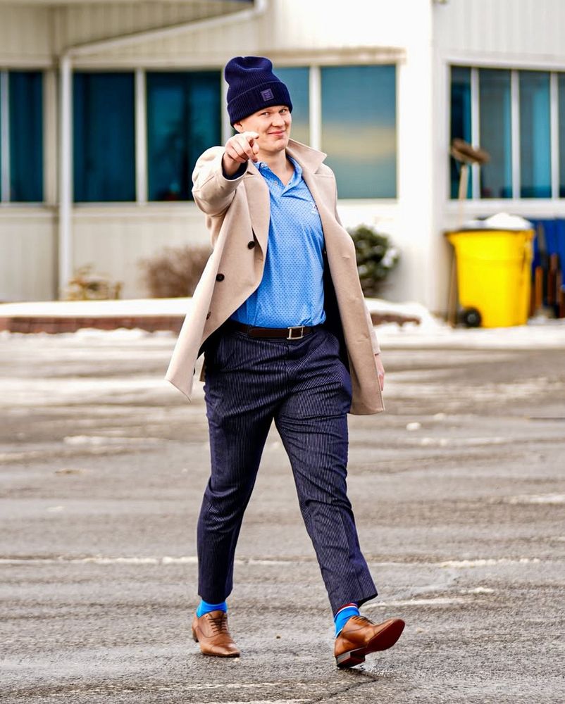 Brady Tkachuk points toward the camera with a smirk as he walks to the right. He’s wearing a light blue unbuttoned-polo, dark striped pants with a belt, a beige jacket, brown leather shoes, blue socks, and a black beanie that’s loose at the top of his head. A building with white siding and large windows is in the background. 