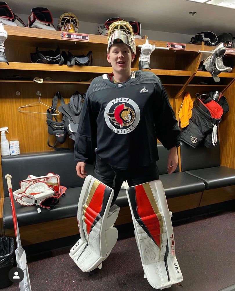 Brady Tkachuk stands in a locker room wearing a black Sens practice jersey; white goalie pads with gold, red, and black stripes on the inside, and a goalie helmet with the mask above his face. He’s standing in front of Anton Forsberg’s stall, with a goalie stick resting on the seat to the left. Various pieces of hockey equipment are on the shelf above his head and hanging behind him. 